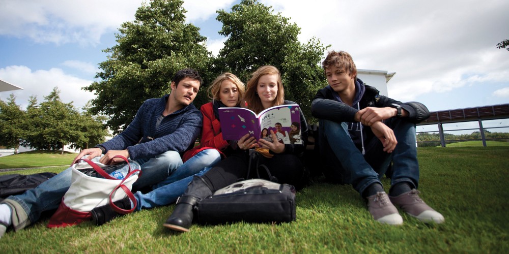 4 students sit on a campus green reading a book together