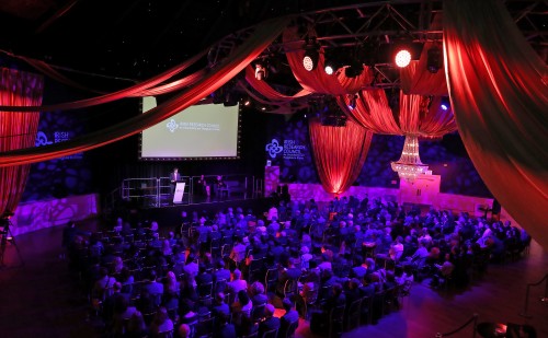 An event hall with red drapes on the ceiling and purple lighting filled with a seated audience, a stage, with a projector in the background