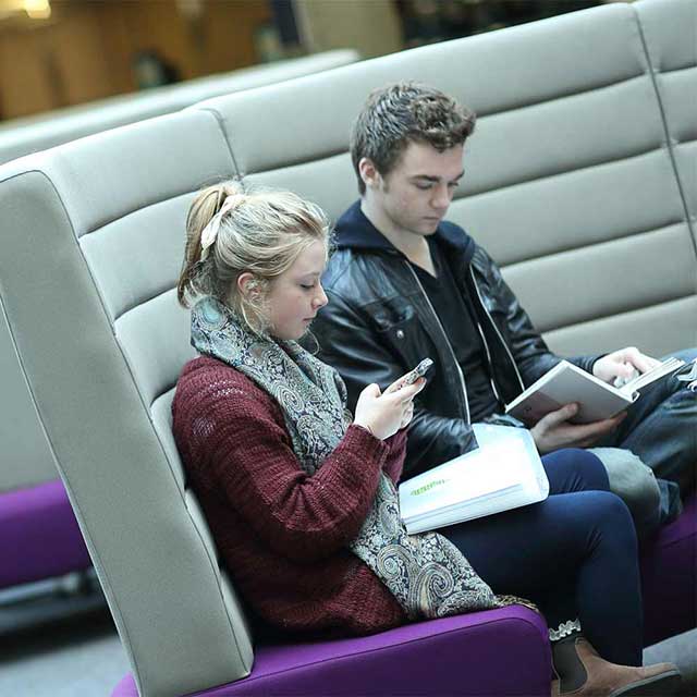 Two students sit on a sofa on campus with a book on their lap