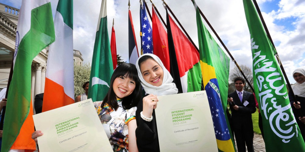 Two students pose smiling with certificates in front of various flags