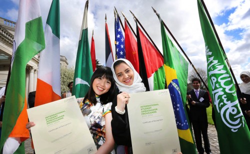 Two students pose smiling with certificates in front of various flags