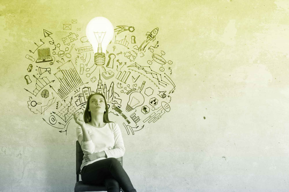 Woman sits in a chair in front of a wall covered in illustrations. She looks up at a lightbulb above her.