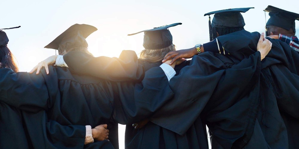 Students with their arms around each others shoulders looking into the sun wearing graduation gowns and caps