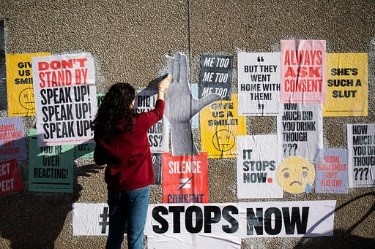 A woman stands in front of a wall covered in posters about consent as she pastes more posters up.