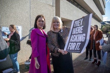 Two women stand in front of a building holding a sign that reads: They were asking for it.