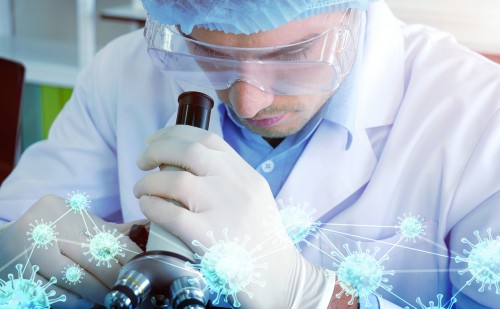 A man with safety glasses and rubber gloves and a lab coat looking through a microscope