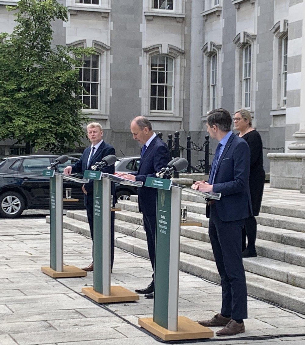 Mícheal Martin, Tim Conlon, and Simon Harris stand at 3 seperate government podiums making an announcment. A woman stands behind them.