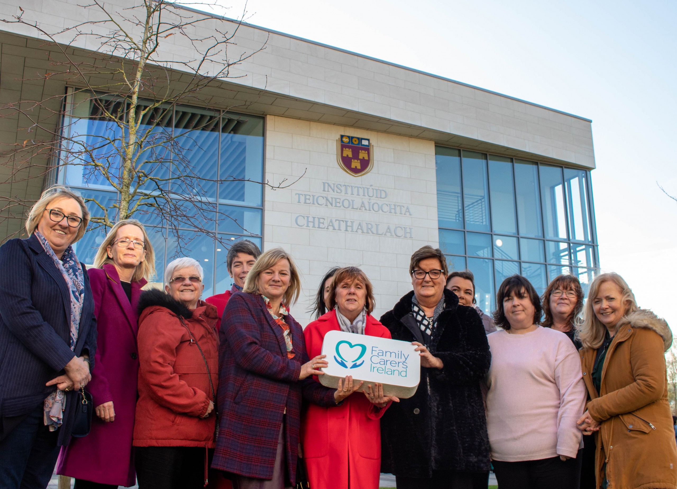 A group photo in front of an IT Carlow campus building with aa group of people holding a sign with the Family Carers Ireland Logo.