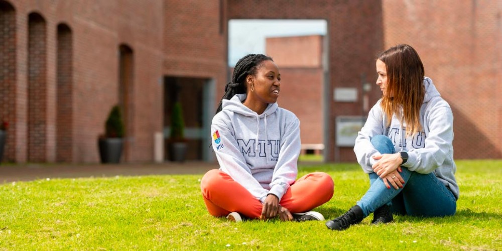 Two Munster Technological University students sit chatting on the grass on campus in front of a red brick building. Both wear grey Munster University hoodies.