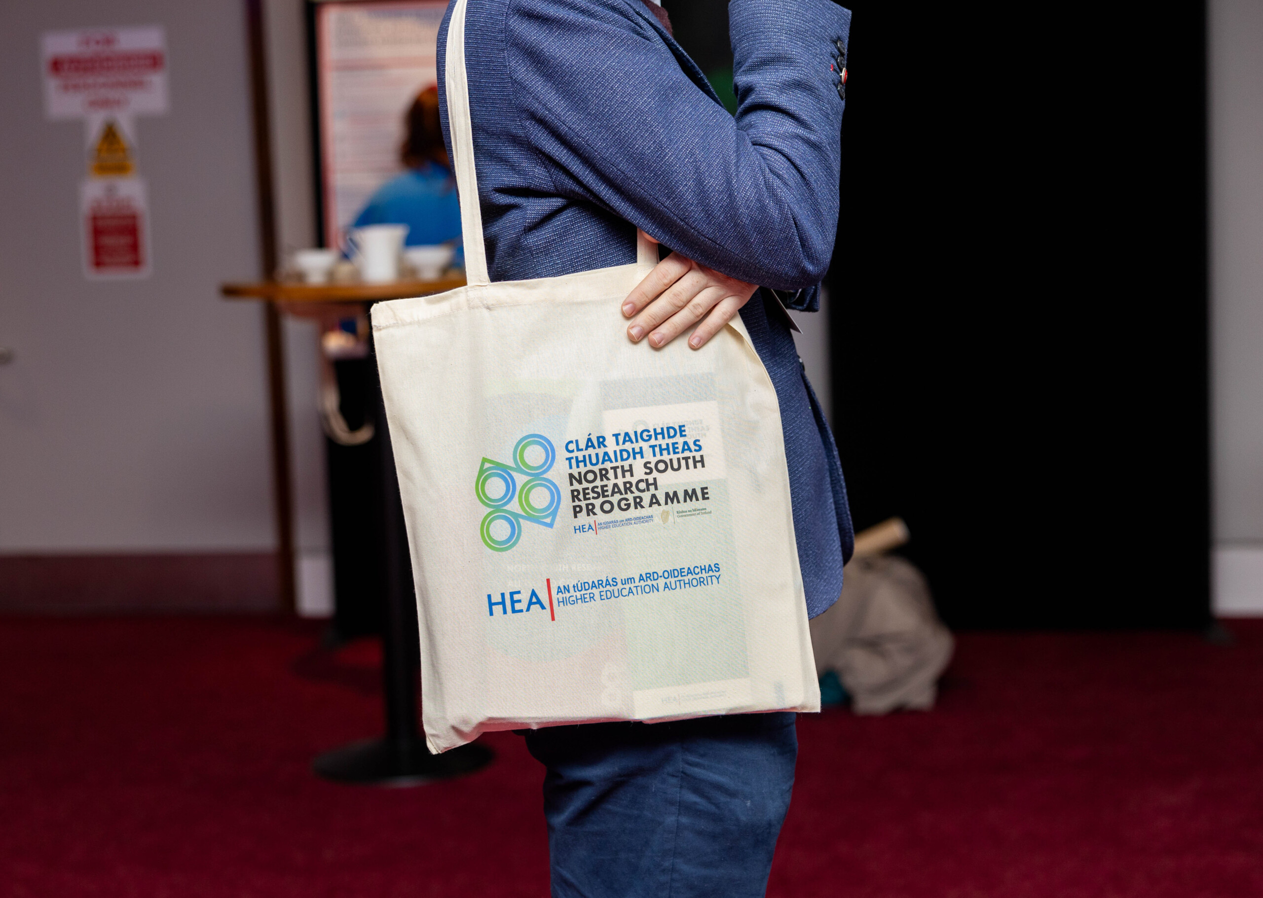 Photo of a person from the shoulders down wearing a tote bag with the North South Research Programme and Higher Education logo on it.