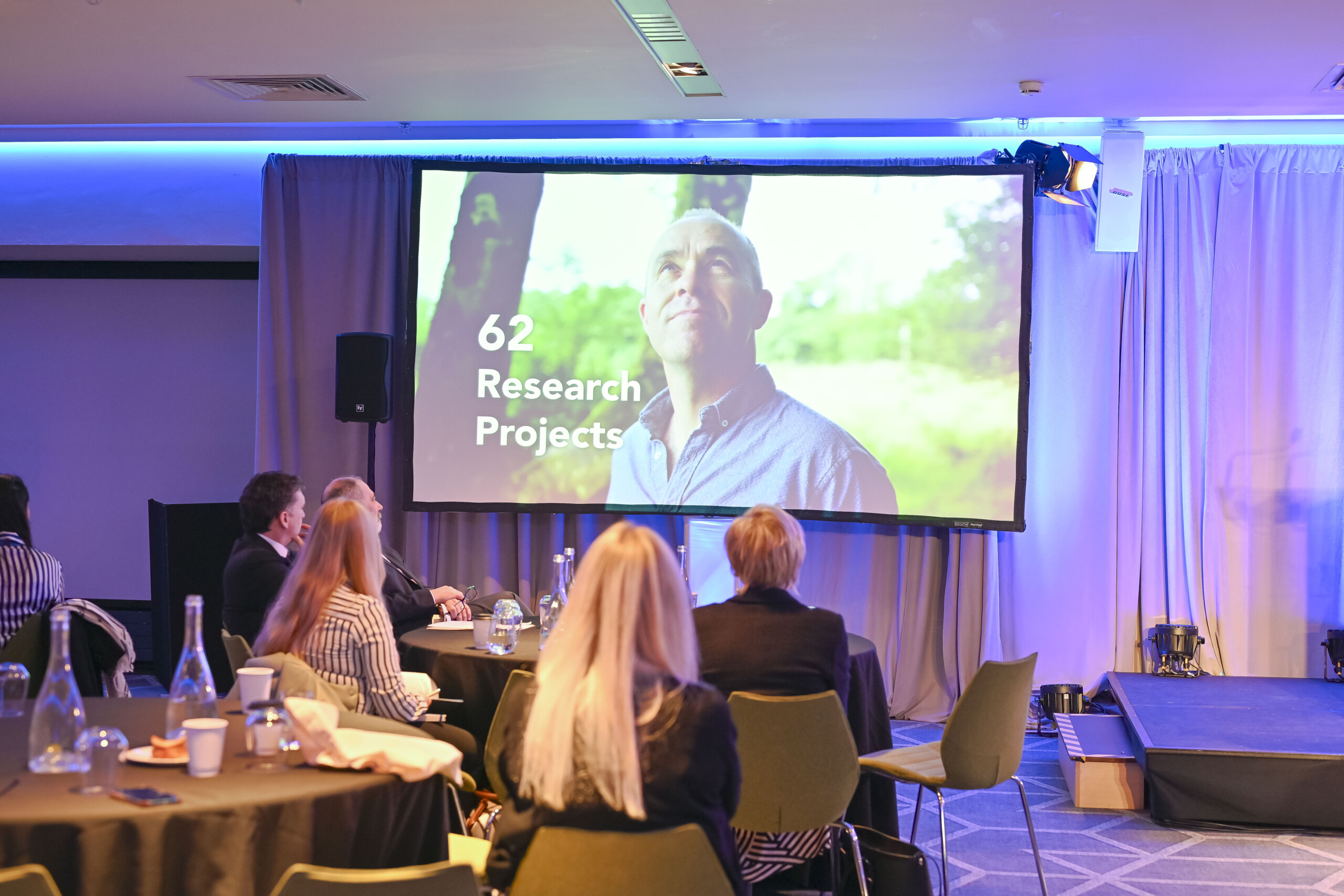 Image of a group of people sitting around a round table watching a presentation on a screen at the North South Research Conference 2024 in Croke Park, Dublin