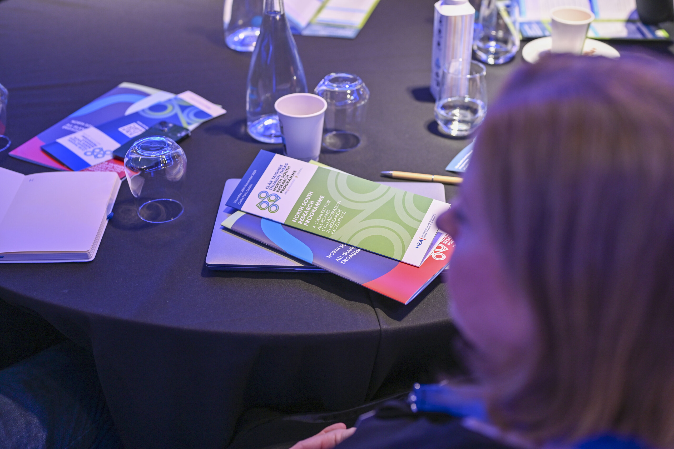 Image of a blurred face in front of a table on which there are information booklets. Taken at the North South Research Programme Conference 2024 at Croke Park, Dublin.