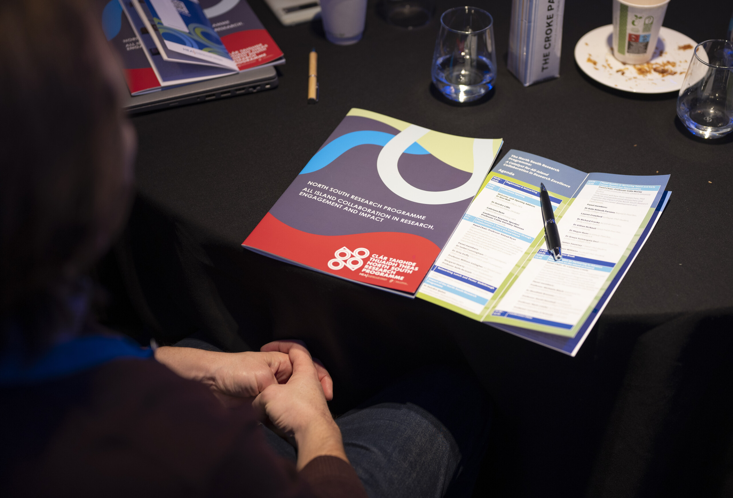 Image of a blurred face with crossed hands in front of a table on which there are information booklets. Taken at the North South Research Programme Conference 2024 at Croke Park, Dublin.