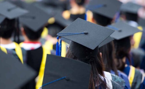 Group of graduates wearing caps and gowns at a graduation ceremony, viewed from behind.