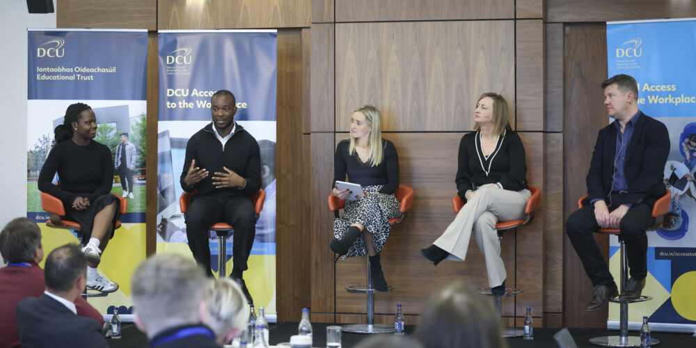 Group of speakers seated on a small platform speaking to an audience